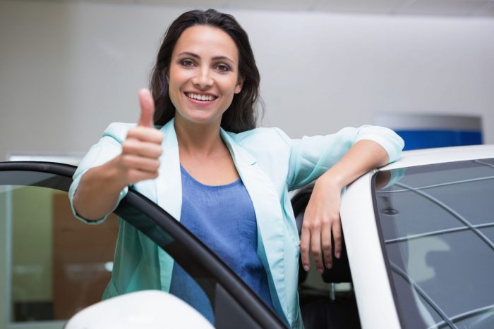 Photo of smiling woman giving the thumbs up leaning on a car illustrates blog: "Looking for Low-Cost Auto Insurance in Texas? Use These Tips"