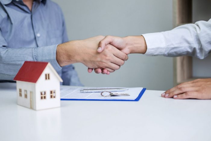 Photo of two men shaking hands next to a scale model of a house illustrates blog: "Are You Required to Have Homeowners Insurance in Texas?"